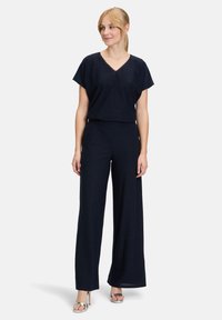 Marineblaues Zweiteiler-Outfit mit einem lässigen V-Ausschnitt-Oberteil und weit geschnittenen Hosen. Der Stoff hat eine weiche Textur mit dezentem Glanz. Silberne Absätze vervollständigen den Look.