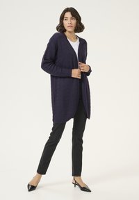 Langärmlige, offen gestaltete Strickjacke in dunkelviolett mit strukturiertem Muster. Kombiniert mit figurbetonten schwarzen Hosen und spitzen Absätzen.