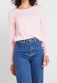 Suéter de manga larga rosa claro, hecho de material suave, combinado con vaqueros azules de talle alto que tienen un cierre de botón y bolsillos frontales.