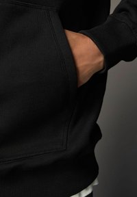 Main dans la poche avant d'un sweat à capuche noir, montrant des coutures détaillées et un poignet à côtes contre un fond gris.