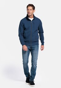 Marineblauwe zip-up jas van textuurstof, met staande kraag en een borstzak, gecombineerd met lichtblauwe jeans en zwarte schoenen.
