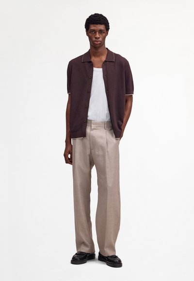 Junger Mann steht und trägt ein dunkelblaues, kurzärmliges Hemd mit Knöpfen, ein weißes Unterhemd, beigefarbene Hosen und schwarze Schuhe vor einem einfarbigen weißen Hintergrund.