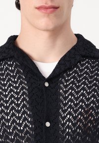 Camisa de punto abierto de color azul marino con cuello, que presenta un patrón en zigzag, cierre de botones y está superpuesta a una camiseta blanca.