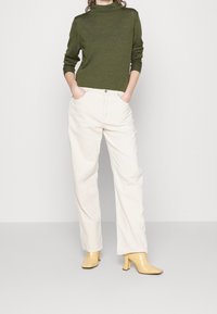 Woman wearing olive green turtleneck sweater, off-white straight-leg pants, and beige heeled ankle boots, standing with hands in pockets.