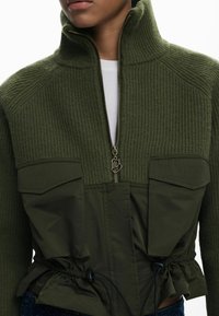 Giacca a zip verde oliva con texture a coste nella parte superiore, tasche piatte e vita regolabile con lacci. Dettaglio della zip in oro.