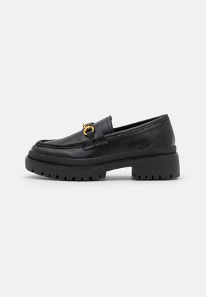 Mocassin en cuir noir avec semelle en caoutchouc épaisse et détail en métal doré en forme de mors, vue latérale sur fond clair uni.