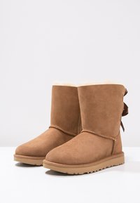 Botas de tobillo de ante marrón con un suave forro de piel de oveja color crema, que presentan una punta redondeada y un lazo decorativo en la parte trasera. Suela de goma plana.
