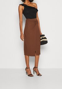 Svart enaxlad bodysuit med volanger, kombinerad med en brun midikjol med rynkor och en sidoklyvning, accessoriserad med en svart och krämrandig väska.