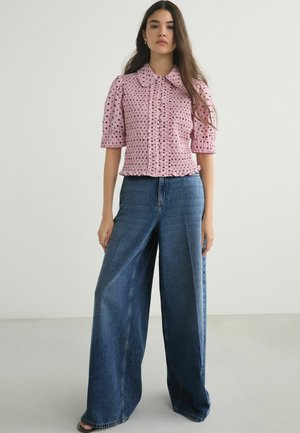 Kvinde iført en lyserød bluse med korte ærmer med sorte polkaprikker og blå denimbukser med vide ben står mod en ensfarvet baggrund.
