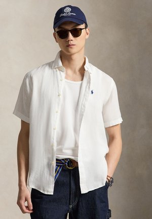 Nuori mies, jolla tummat aurinkolasit, tummansininen Ralph Lauren -lippis, valkoinen lyhythihainen nappisärkkä valkoisen T-paidan päällä, tummat farkut raidallisella vyöllä.