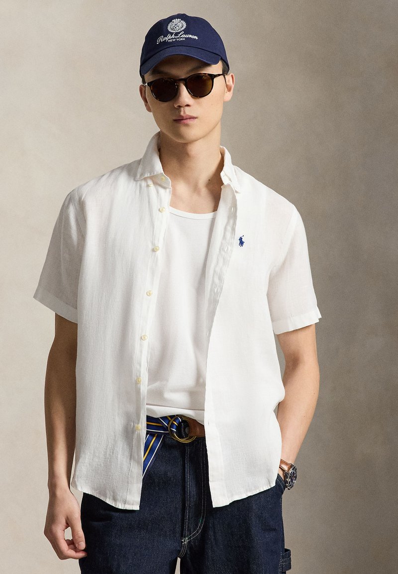 Mladý muž s tmavými slnečnými okuliarmi, tmavomodrá čiapka Ralph Lauren, biela krátkorukávová košeľa zapnutá na gombíky cez biele tričko, tmavé džínsy s pruhovaným opaskom.