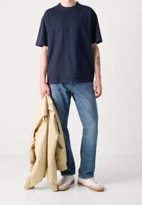 Mörkblå bomullströja med rund halsringning, i kombination med ljusblå denimjeans och en beige bomberjacka som hålls i ena handen. Vita sneakers.