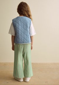 Gepolsterte blaue Weste, weißes T-Shirt, grüne gestreifte weit geschnittene Hose und weiße Sneaker. Rückansicht, heller Holzboden, neutraler Hintergrund.