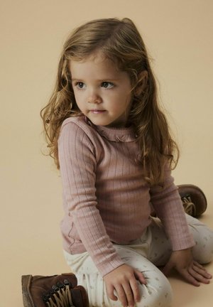 Maglione rosa a righe con maniche lunghe e scollo a volant, pantaloni crema a fantasia e stivali marroni con lacci. Sfondo marrone chiaro.