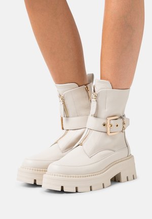 Bottes blanches en cuir épaisses à la cheville avec double fermeture éclair, une large sangle avec une boucle dorée, et des semelles plateformes épaisses et texturées couleur crème.