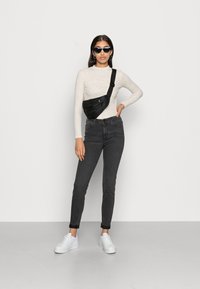 A mala de ombro preta apresenta uma alça única e um bolso com fecho. O modelo usa uma blusa clara com manchas, jeans escuros e ténis brancos.