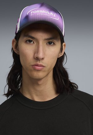Młoda osoba z długimi ciemnymi włosami, nosząca fioletową czapkę "Formula 1 Racing League" i czarną koszulkę, patrzy przed siebie z neutralnym wyrazem twarzy.
