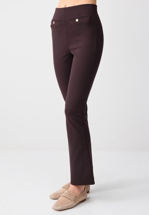 Vrouw draagt slanke, donkerbruine broek met voorzakken met knopen en beige platte loafers, staand op een lichte achtergrond.