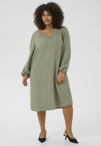 Robe vert olive avec des pois noirs, décolleté en V, manches bouffantes et coupe ample, assortie à des chaussures noires à petits talons.