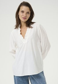 White blouse with a ruffled neckline, long sleeves, and a relaxed fit, made from lightweight fabric. Paired with light blue jeans.