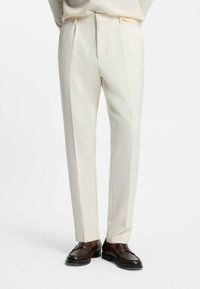 Beige op maat gemaakte broeken met scherpe vouwen, een slim fit, zijzakken en riemlussen, gecombineerd met bruine loafers. Gladde textuur.
