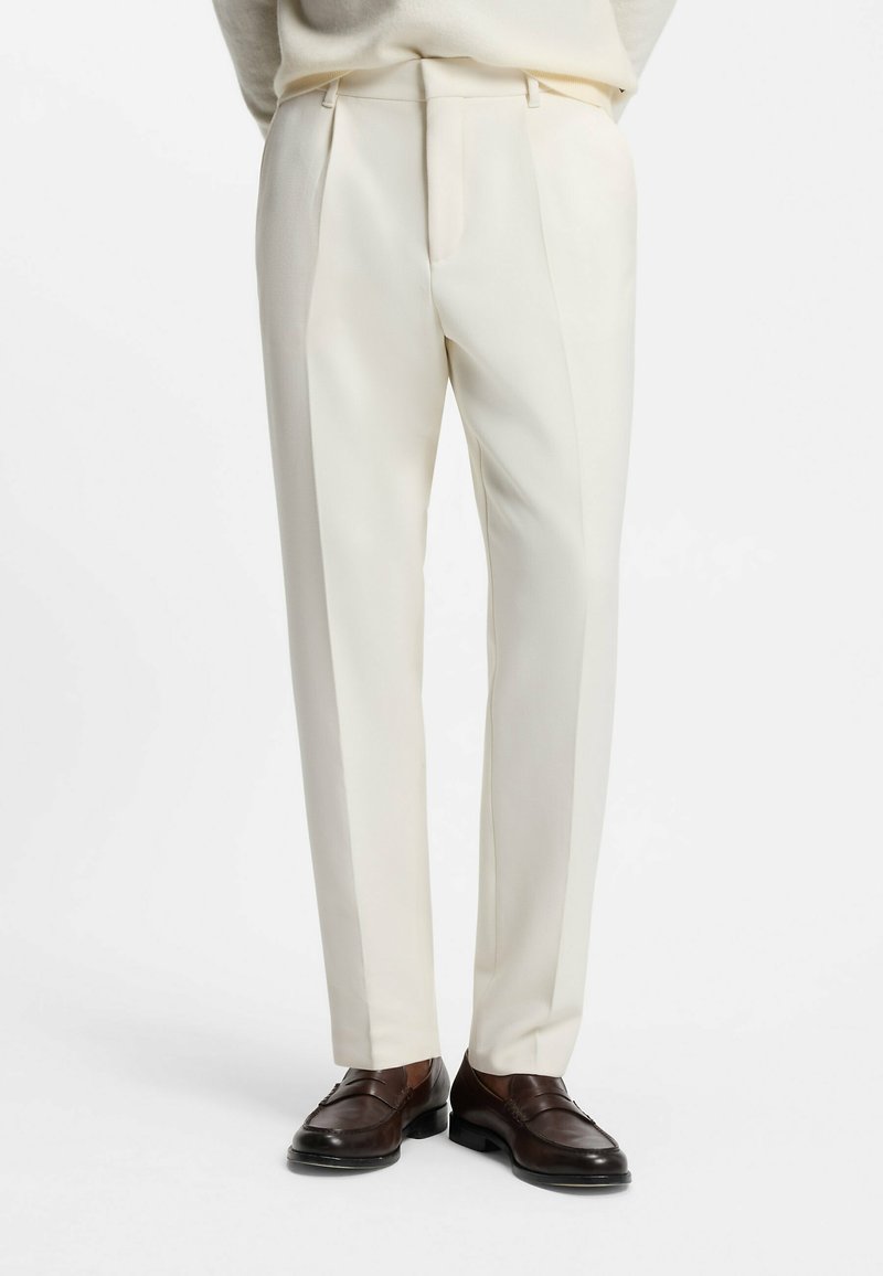 Beige op maat gemaakte broeken met scherpe vouwen, een slim fit, zijzakken en riemlussen, gecombineerd met bruine loafers. Gladde textuur.