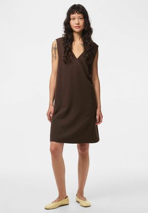 Femme aux longs cheveux foncés, portant une robe marron sans manches jusqu'aux genoux et des chaussures plates beiges, debout sur un fond blanc uni.