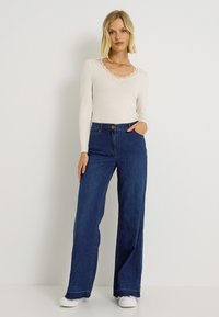 Blusa de manga larga color crema con encaje en los bordes, combinada con jeans de pierna ancha en azul oscuro que presentan un bolsillo frontal y detalles de botón. Zapatillas completan el look.