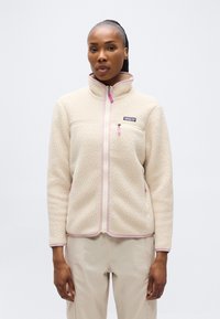 Γυναίκα που φοράει κρεμ φλις μπουφάν με ροζ φερμουάρ και ανοιχτό μπεζ παντελόνι, στέκεται μπροστά σε απλό λευκό φόντο.