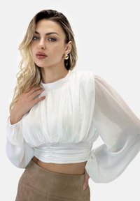 Hvid bluse med gennemsigtige lange ærmer, rynket krop og glat tekstur. Båret med en tan nederdel. Guldøreringe tilføjer et kontrasterende element.