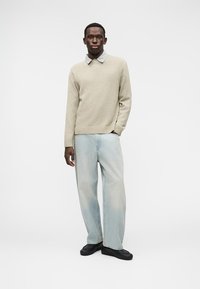 Πλεκτό μπεζ πουλόβερ πάνω από ανοιχτό γαλάζιο πουκάμισο με γιακά, συνδυασμένο με ξεθωριασμένο γαλάζιο τζιν με φαρδιά μπατζάκια και μαύρα παπούτσια, σε μινιμαλιστικό φόντο.