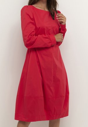 Kobieta ubrana w czerwoną sukienkę na całe ręce, do kolan, z bufkami na ramionach i marszczonymi mankietami, stojąca na tle jednolitego jasnego tła.