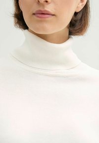 Záběr zblízka na osobu, která má na sobě bílý žebrovaný rolák, vidět je krk, brada a část rtů.