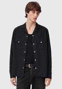 Sort skjorte med broderede detaljer, forlommer, sølvaccents og knaplukning. Båret over en sort skjorte, kombineret med mørke jeans.