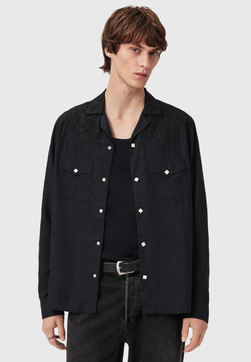 Sort skjorte med broderede detaljer, forlommer, sølvaccents og knaplukning. Båret over en sort skjorte, kombineret med mørke jeans.