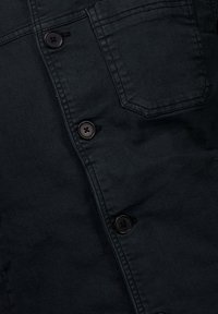 Camisa de mezclilla negra con grandes botones negros, un bolsillo en el pecho y una textura suave. Tiene una silueta estructurada con costuras visibles.