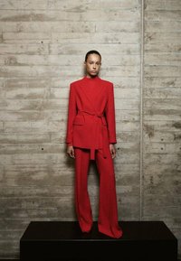 Woman standing on black platform wearing a red belted blazer and matching wide-leg trousers against a textured concrete wall.