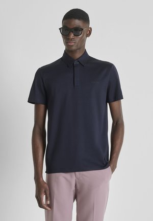 Man die zwarte zonnebril draagt, donkerblauw poloshirt met korte mouwen en lichtroze broek, staand met één hand in de zak.