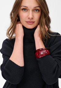 Zwarte coltrui met ribbeltextuur, met drie armbanden: twee glanzende rode en één donker tortoise. 