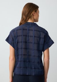 Marineblaue kurz�ärmelige Bluse mit einem strukturierten Rautenmuster. Der Stoff wirkt leicht und semi-transparent, mit einem geraden Saum und rundem Ausschnitt.