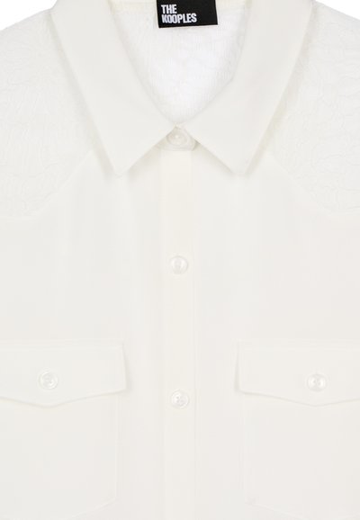 Camicia bianca con bottoni, caratterizzata da spalle in pizzo testurizzato, due tasche frontali e bottoni perlacei. Colletto sottile con finitura in tessuto liscio.