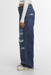 Personne portant un pantalon denim bleu déchiré taille haute et des baskets blanches avec le texte "GAME SET MATCH" sur la semelle, associé à un haut blanc à manches longues.
