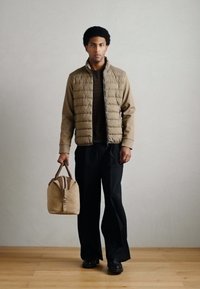 Veste matelassée marron portée sur une chemise foncée, associée à un pantalon noir à jambes larges. Tenant un sac beige avec des accents marron, debout sur un sol en bois.