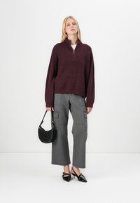 Un pull à fermeture éclair bordeaux, un pantalon cargo gris, un sac à main noir clouté et des talons aiguilles noirs, présentant une tenue décontractée et casual.