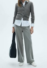 Grijze cardigan met knopen over een wit shirt, gecombineerd met brede grijze broek en een zwarte schoudertas. Witte sneakers maken de look compleet.