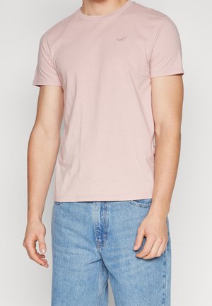Mann trägt ein hellrosa kurzärmliges T-Shirt und hellblaue Jeans, steht vor einem neutralen Hintergrund.