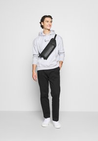 Sorte crossbody taske med tekstureret overflade, båret over en lys grå hoodie, parret med sorte bukser og hvide sneakers. Simpelt design.