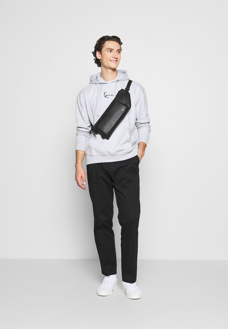 Sorte crossbody taske med tekstureret overflade, båret over en lys grå hoodie, parret med sorte bukser og hvide sneakers. Simpelt design.