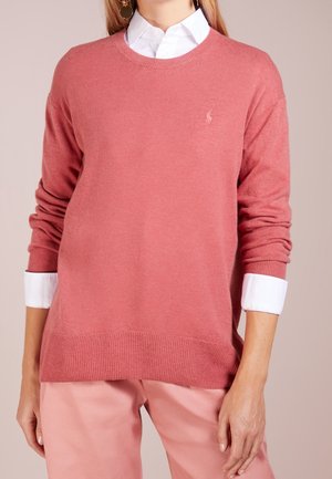 Femme portant un pull rose clair à col rond par-dessus une chemise blanche à col avec un pantalon rose, vue de la poitrine jusqu'aux hanches.