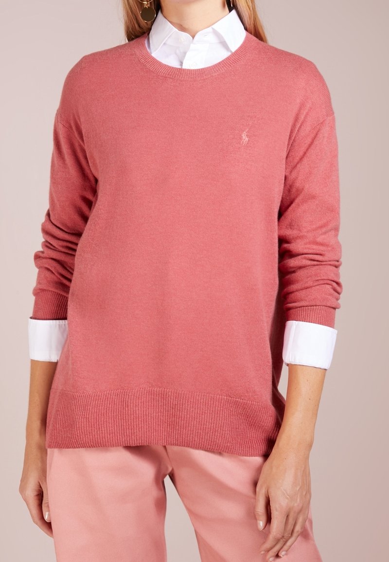 Femme portant un pull rose clair à col rond par-dessus une chemise blanche à col avec un pantalon rose, vue de la poitrine jusqu'aux hanches.